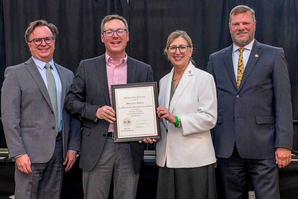 Four people standing together in a row (three men and one woman). The woman and the man in the center are holding a plaque that is for the William J. Beal Outstanding Faculty Award.