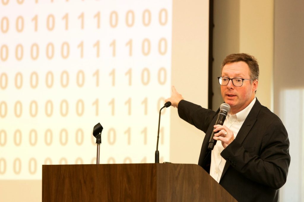 Man standing at a podium with a microphone in his hand pointing at a screen with a series of numbers, all zeros and ones. 