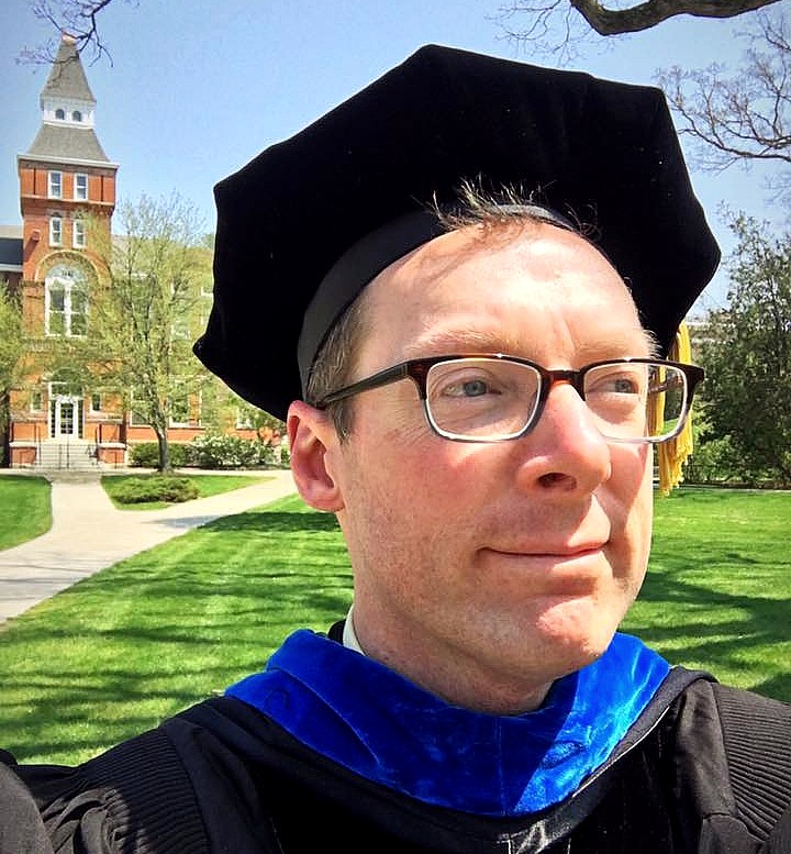 Man wearing a black graduation cap and gown taking a selfie photo of himself with Linton Hall in the background.
