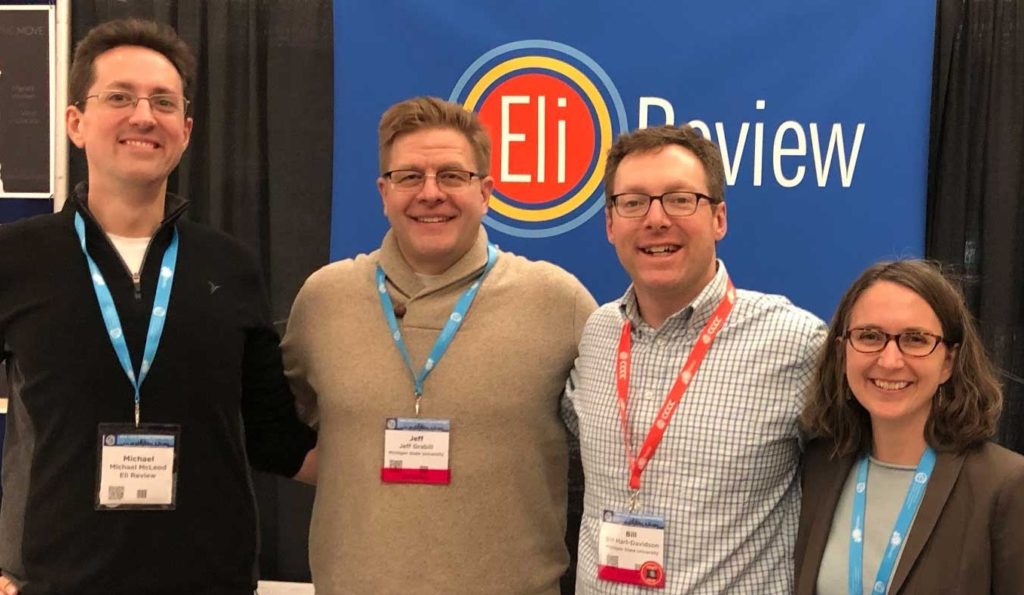 Four people (three men and one woman) who are all wearing glasses and lanyards around their necks, standing in front of a sign that shows the logo for Eli Review. 