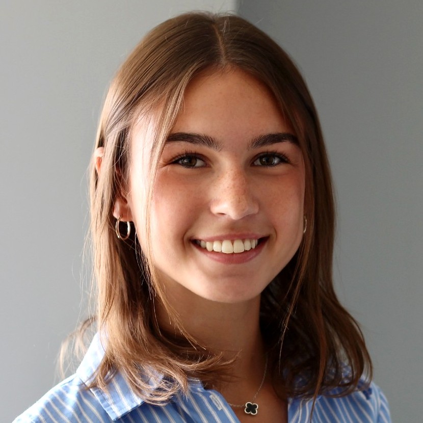 Woman smiling, wearing a blue and white striped button-up shirt, standing in front of a neutral gray background.