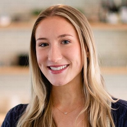 Woman smiling indoors, wearing a dark top and navy cardigan, with light blonde hair and a modern kitchen background.