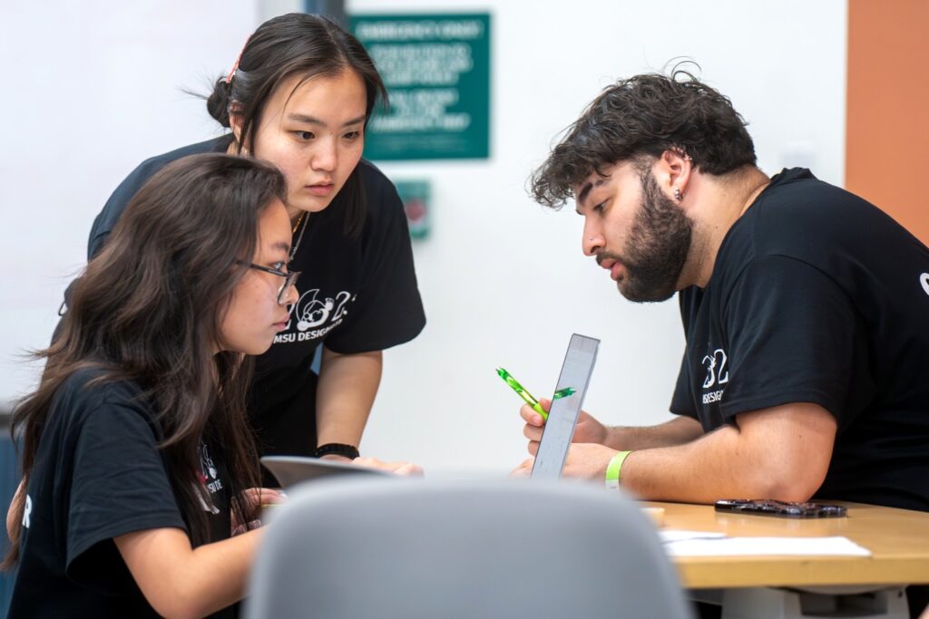 Three students collaborate intently at a table, with one student holding a tablet and pen while the others lean in to view the screen. All are wearing black “MSU Designathon” shirts.
