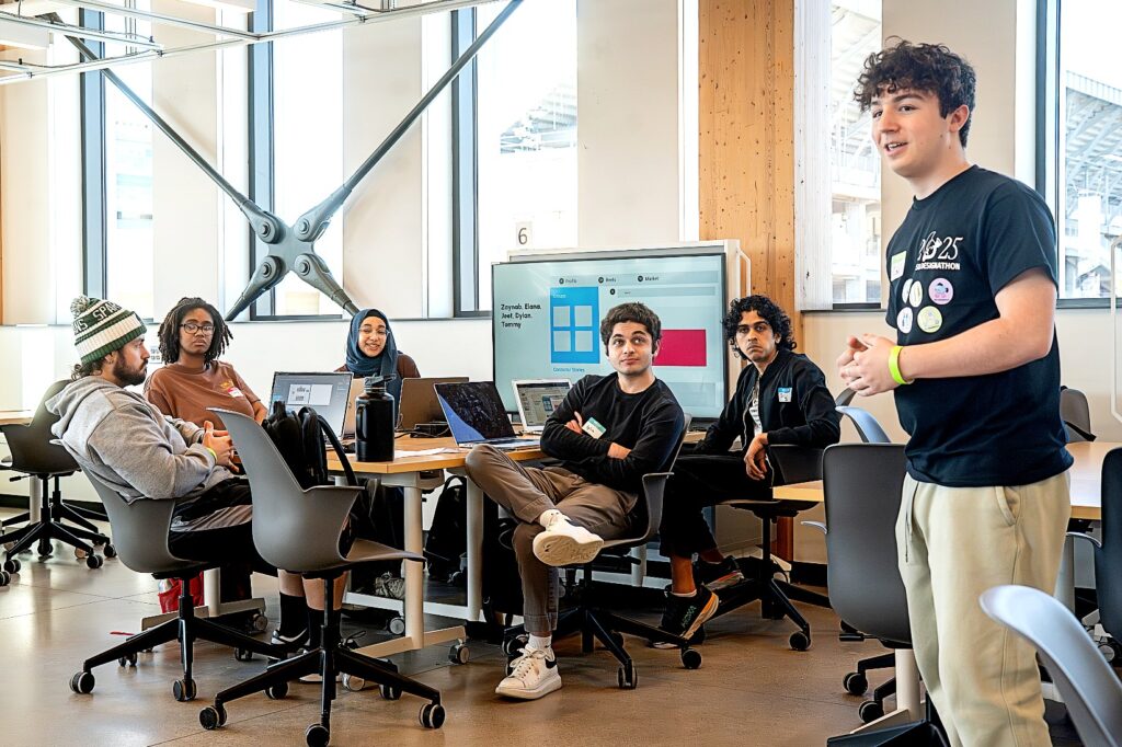 A group of six students engage in a team discussion in a modern classroom, with one student standing and speaking while the rest sit around laptops. A presentation screen is visible in the background.