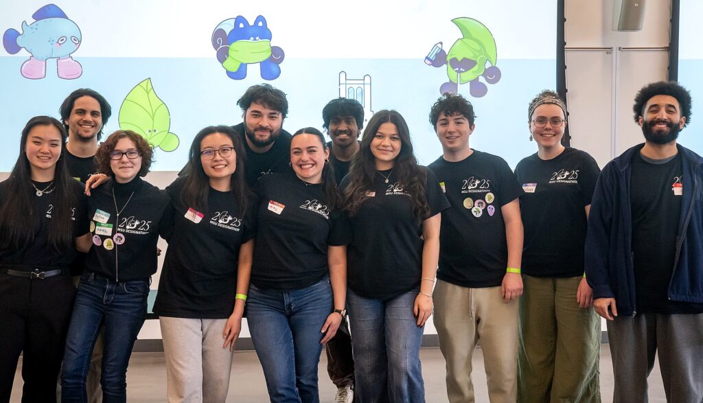 group of eleven students stands side by side, smiling in front of a projection screen displaying colorful cartoon characters. All are wearing matching black t-shirts that read “2025 MSU Designathon,” with name tags and decorative pins.