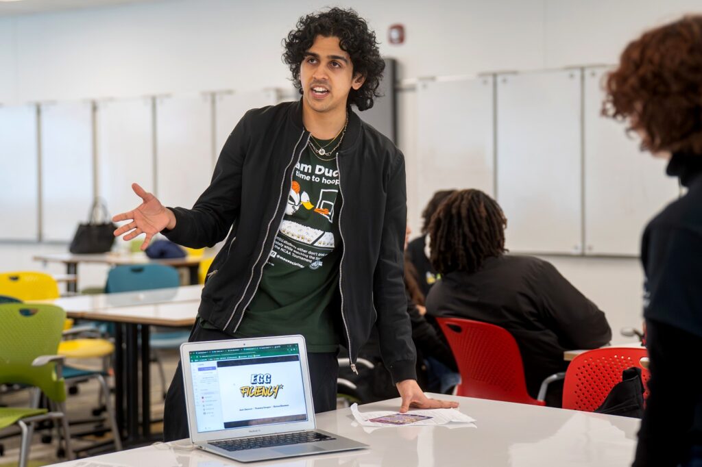 A student presents a project titled “Egg Fluency” using a laptop, gesturing with one hand while standing at a table in front of seated classmates.