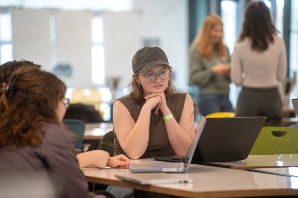 A student in a cap and glasses sits at a table, leaning forward with hands under chin, listening attentively during a group discussion with laptops open.