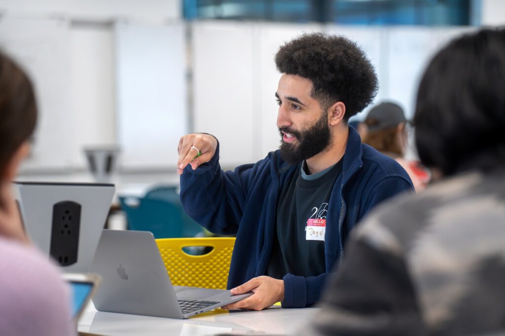 A student with a beard gestures while speaking in a group setting, seated at a table with a laptop. He wears a dark jacket and an “MSU Designathon” shirt.