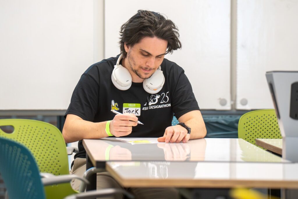 A student sits alone at a table, writing on a name tag labeled “Jack.” He wears headphones around his neck and a black “MSU Designathon” shirt.