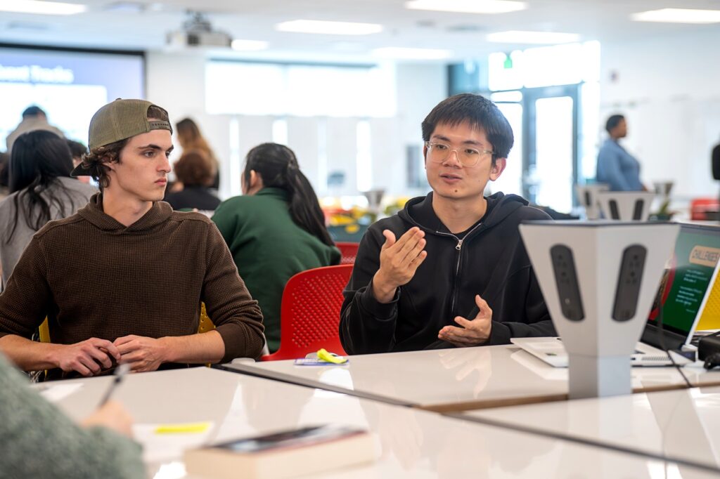Two students sit at a table during a discussion. One, wearing a black zip-up hoodie, gestures with his hands while speaking. The other, in a brown sweater and backwards cap, listens . A laptop and group materials are visible on the table in a well-lit classroom.