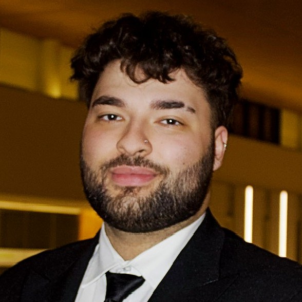 Man dressed in a black suit and tie, posing in a warmly lit indoor space.