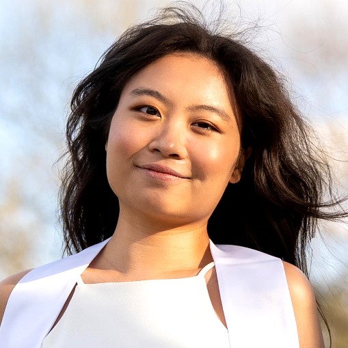 Woman wearing a white graduation stole and white dress, smiling outdoors with trees and soft sunlight in the background.