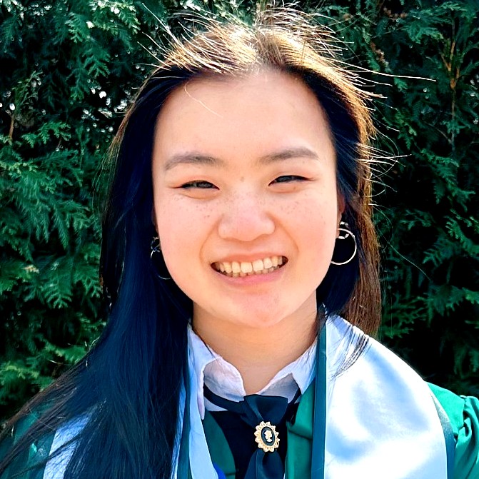 Woman in a green graduation gown with a white stole, smiling in front of a backdrop of evergreen trees