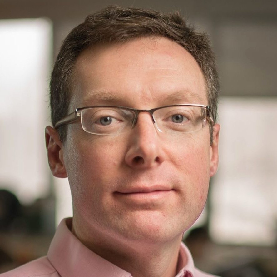 Close-up of a man wearing glasses and a light pink shirt, with a blurred indoor office setting in the background.