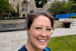 Selfie photo of a woman with dark brown shoulder length hair and glasses, who is smiling and standing outside in front of a stone-colored eight-story buidling.