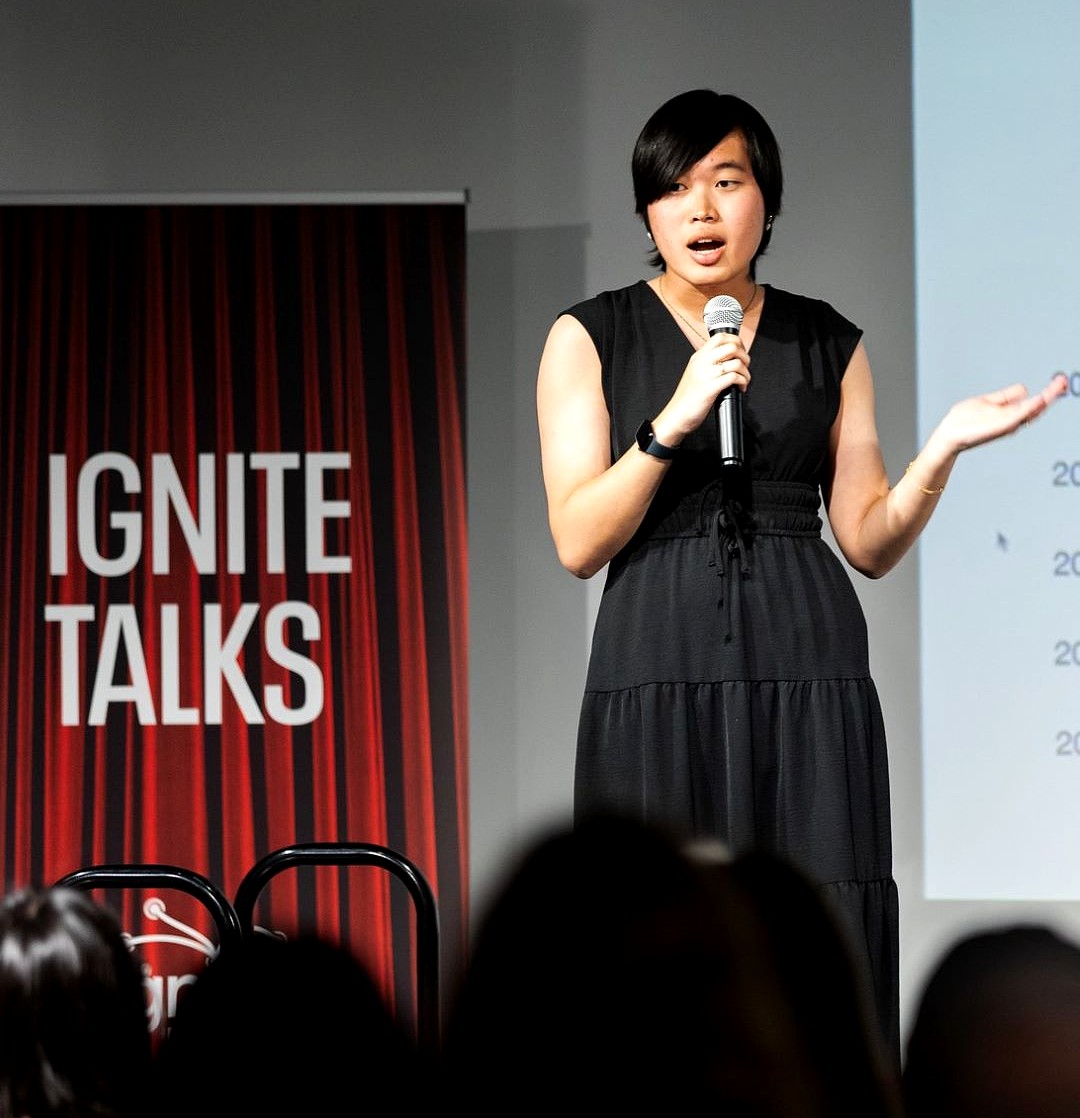 Alex Guo stands on stage at Ignite Talks MSU, speaking into a handheld microphone while gesturing with her left hand. She wears a sleeveless black dress and faces the audience, with an Ignite Talks banner visible behind her.