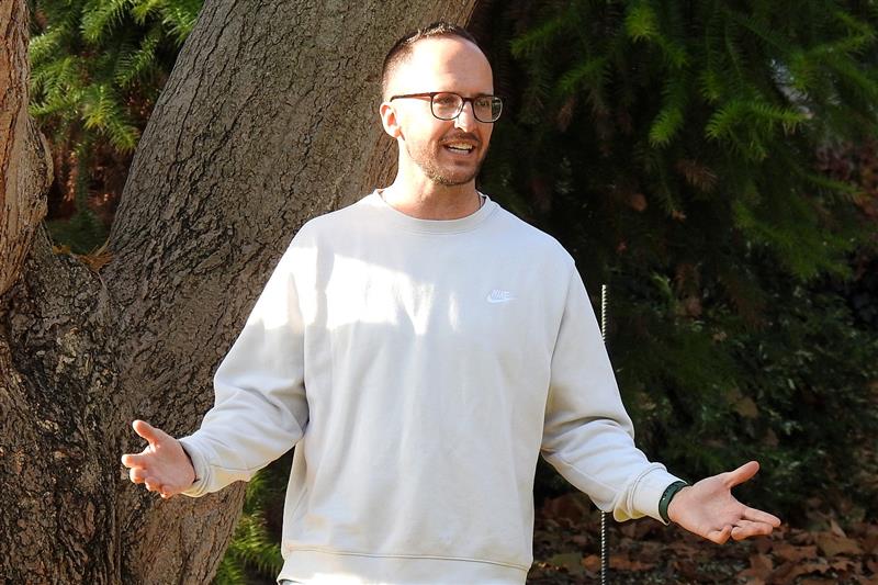 A man standing outdoors near a large tree, wearing a light-colored sweatshirt and glasses, with his hands spread. 
