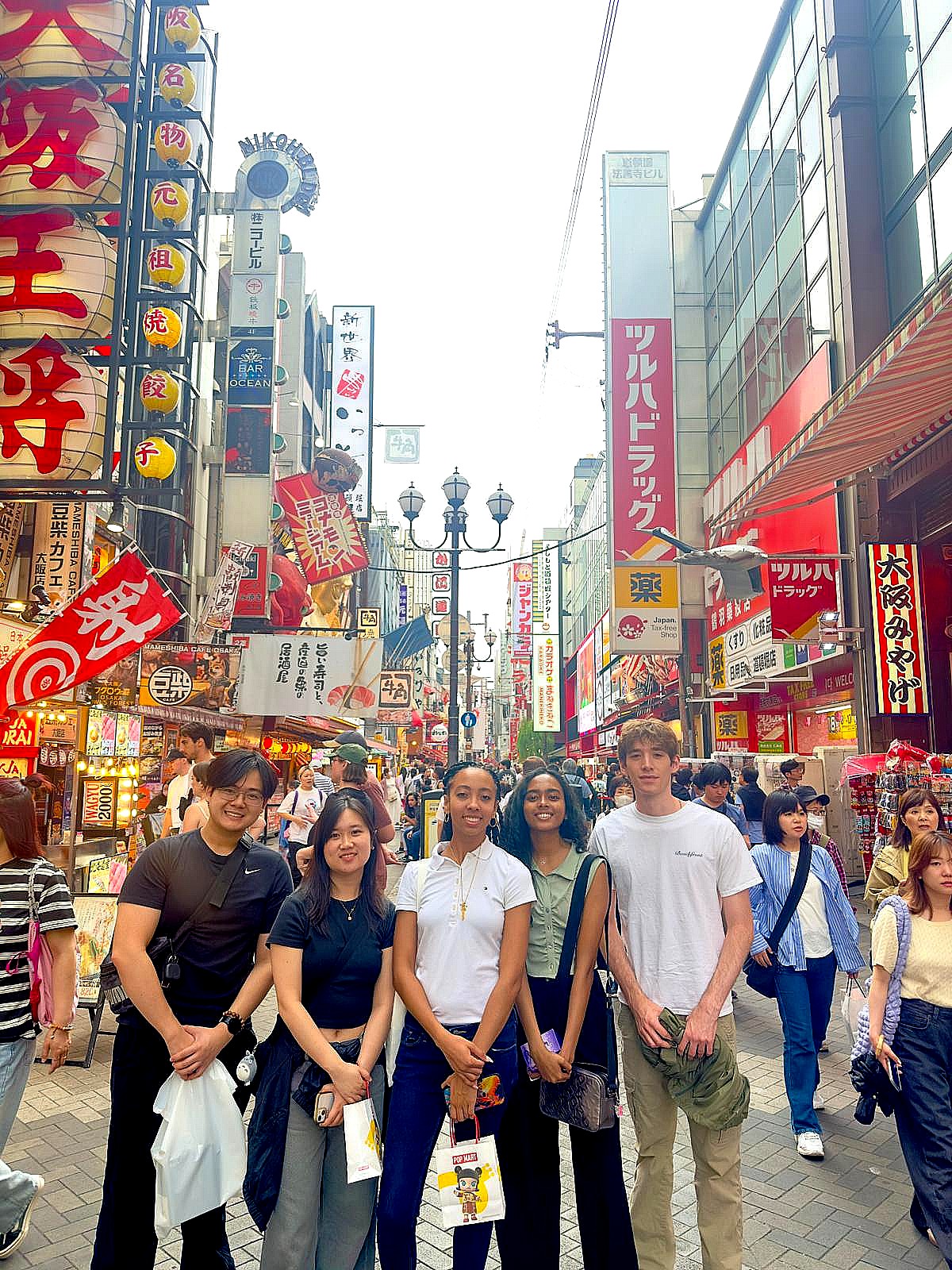 A group of five young adults stands together in the middle of a crowded city street lined with colorful Japanese signs, lanterns, and storefronts, as pedestrians pass by in a lively shopping and dining district.